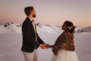 Bride and groom holding hands close to camera out of focus with snow mountains and sunset sky background in focus
