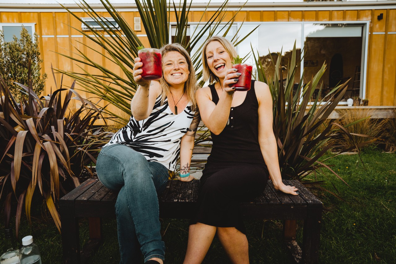 Kellie and Charlotte from Shaken & Stirred Weddings sitting outside in the garden raising their drinks for a cheers
