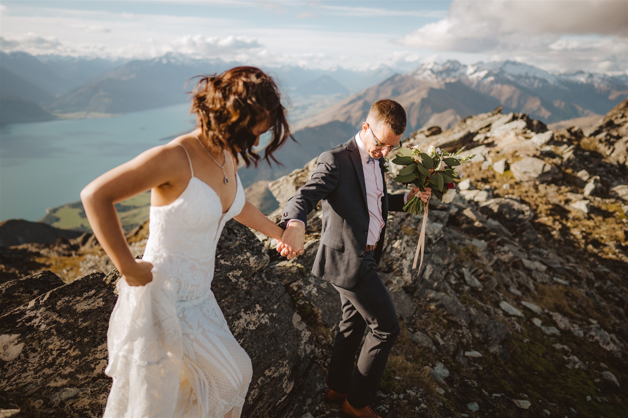 Bride and groom in wedding outfits on mountain top walking over rocky mountains with Glenorchy in the background