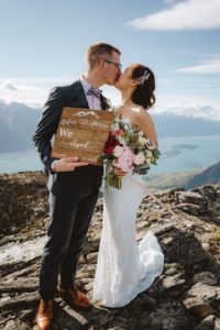 bride and groom holding elopement announcement sign and kissing at Mount Crichton, Queenstown, New Zealand